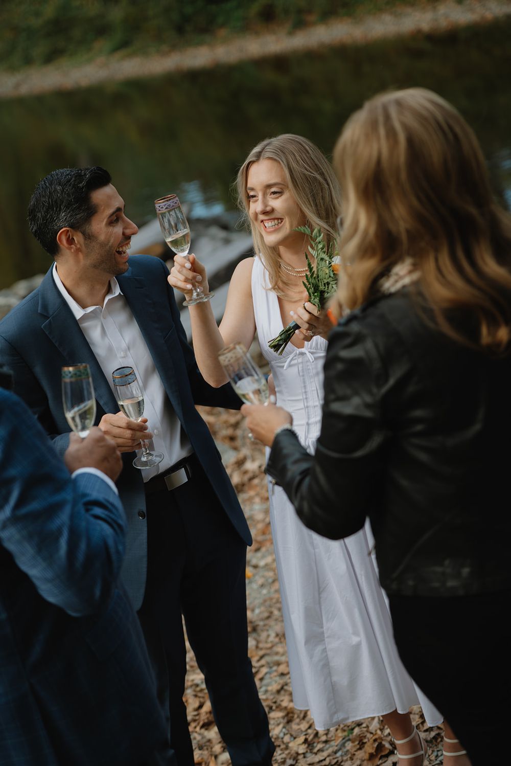 Wedding guests share laughter and champagne during an outdoor celebration by a lake.