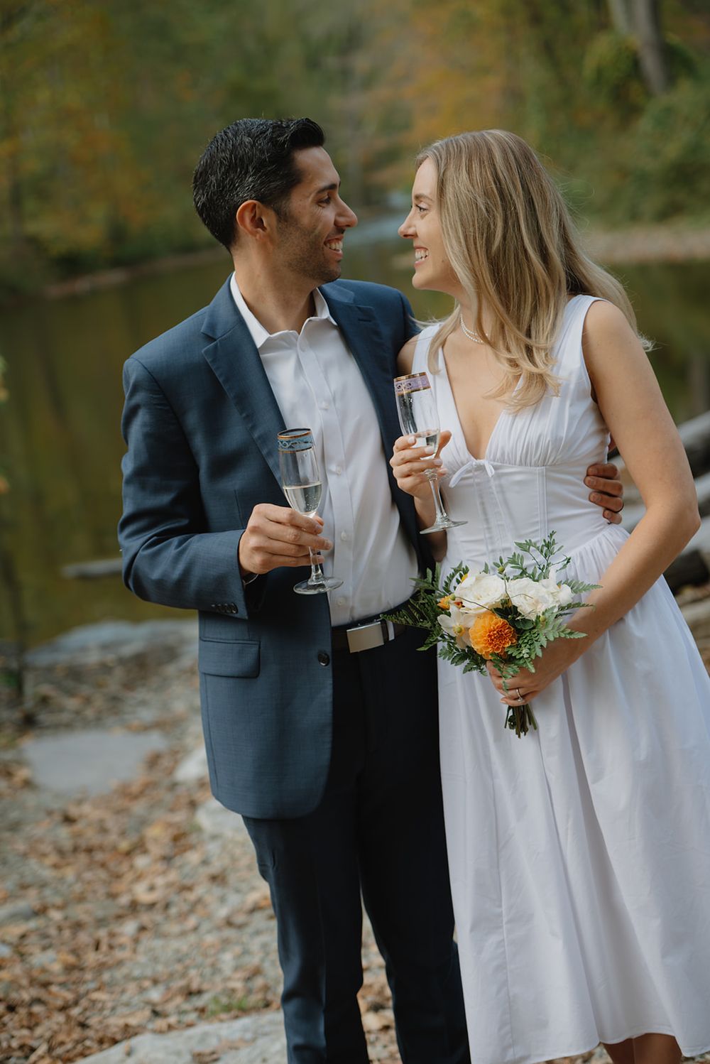 A couple in wedding attire shares a romantic moment with champagne glasses in an autumn forest setting.