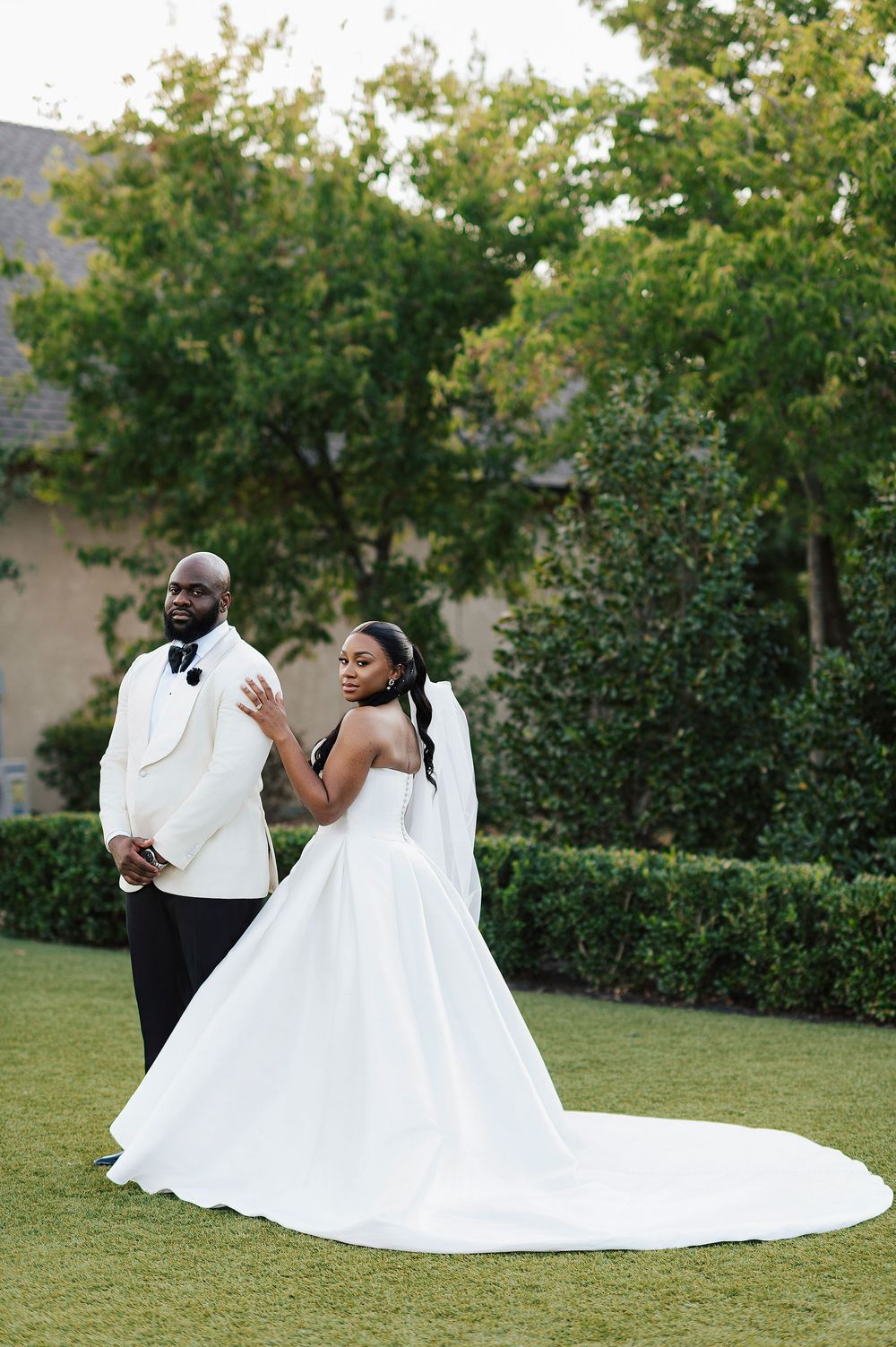 A newlywed couple in formal wedding attire shares a romantic moment on a grassy lawn surrounded by trees.