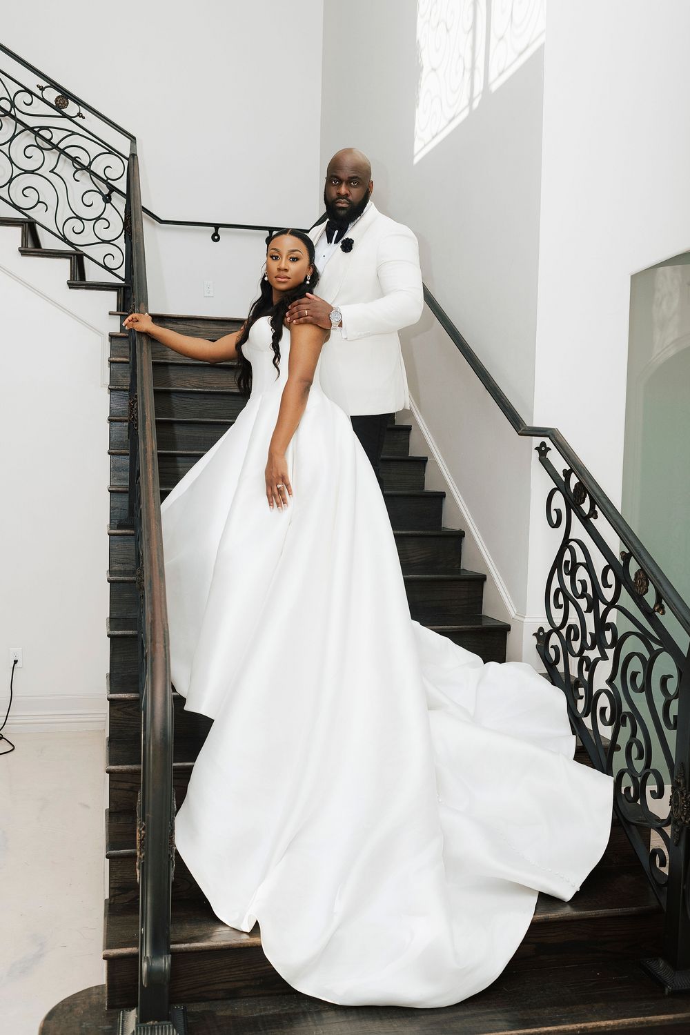 A bride in a flowing white gown poses elegantly on a dark staircase with ornate wrought iron railings.