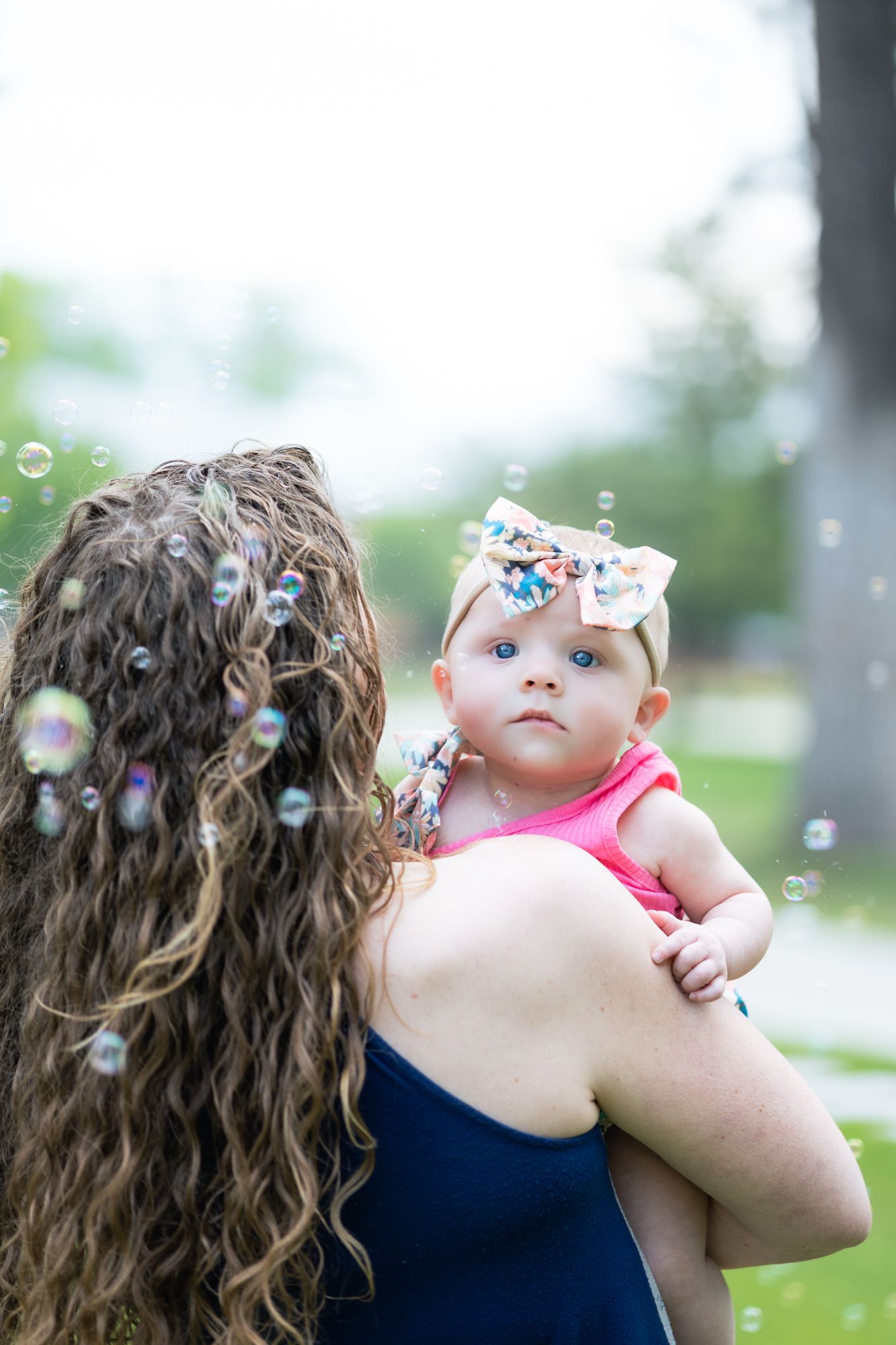 A parent and child blow bubbles together outdoors on a sunny day while wearing navy blue clothing.