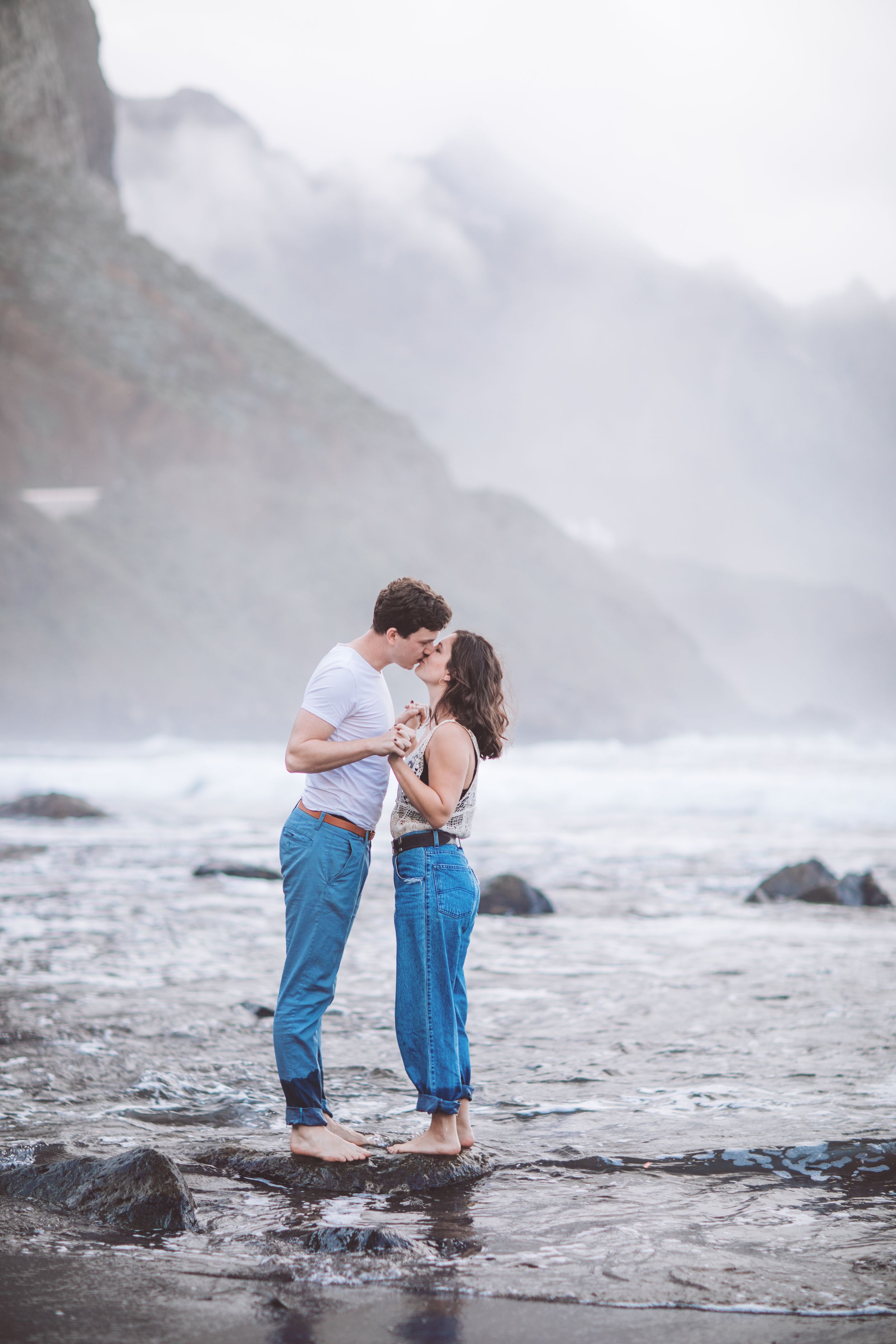 A romantic couple embraces while standing in shallow ocean waters with misty mountains in background.