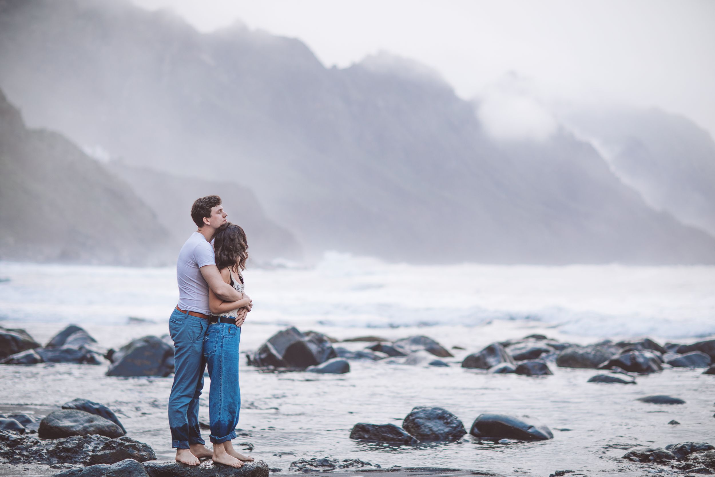 Dramatic oceanside engagement photo with rocky coastline and misty mountains.