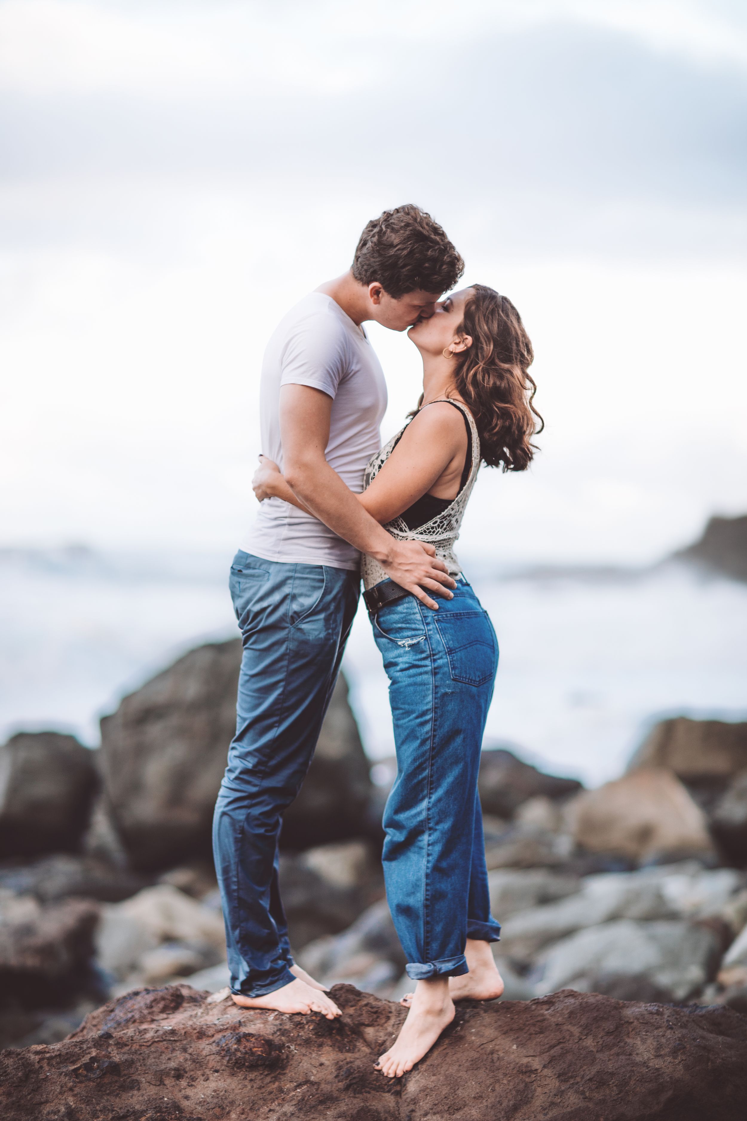 A romantic couple embraces barefoot on coastal rocks during their engagement photoshoot.