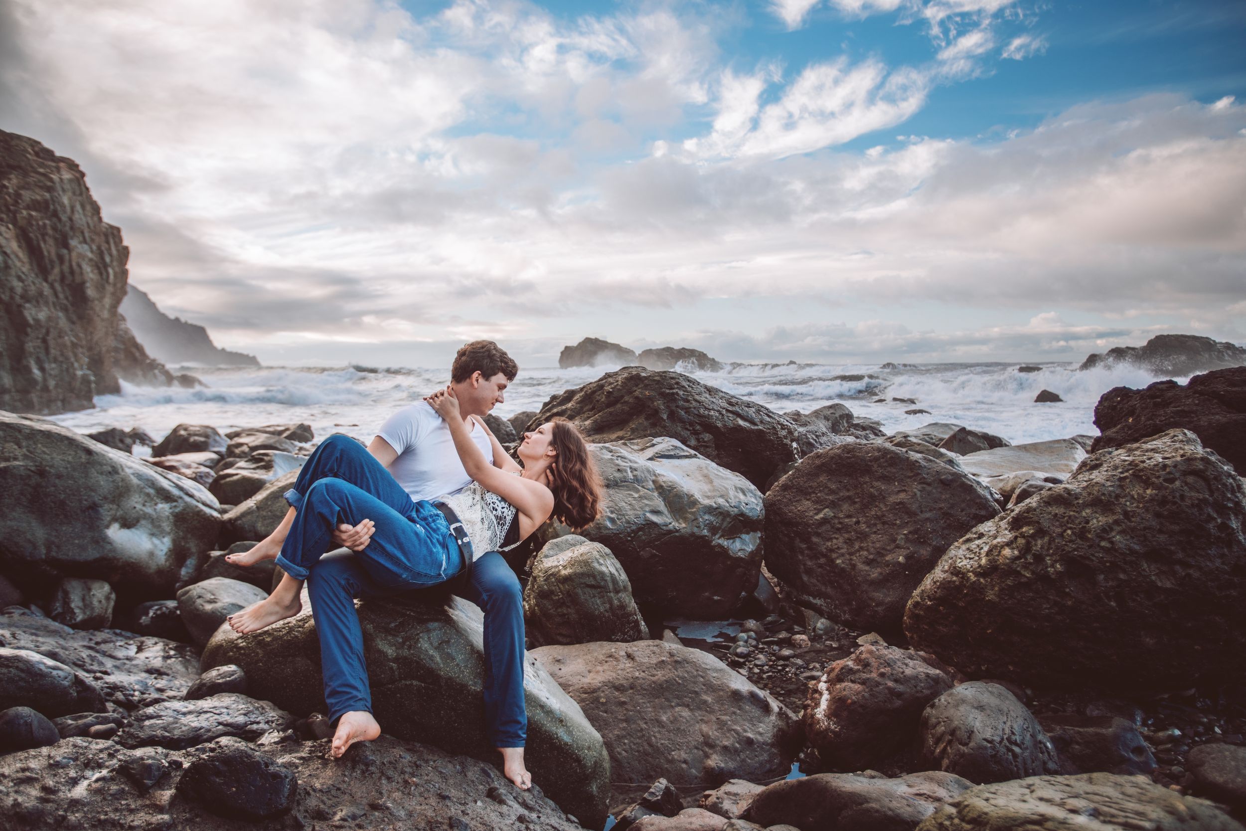 A couple poses on dramatic rocky coastline with crashing waves and cloudy skies.