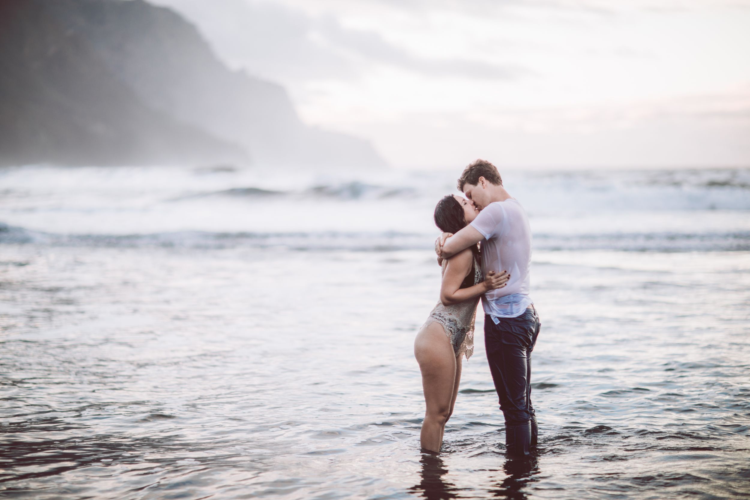 Couple embracing in shallow ocean waves against misty coastal cliffs at sunset.
