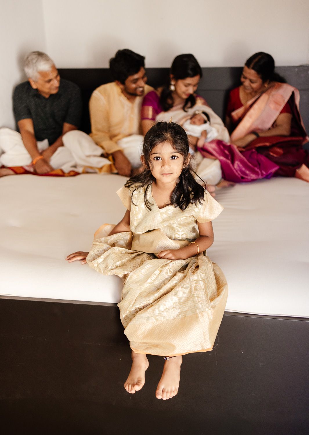 Big sister sits on the bed with her parents and new baby sister in the background during their newborn family photography session