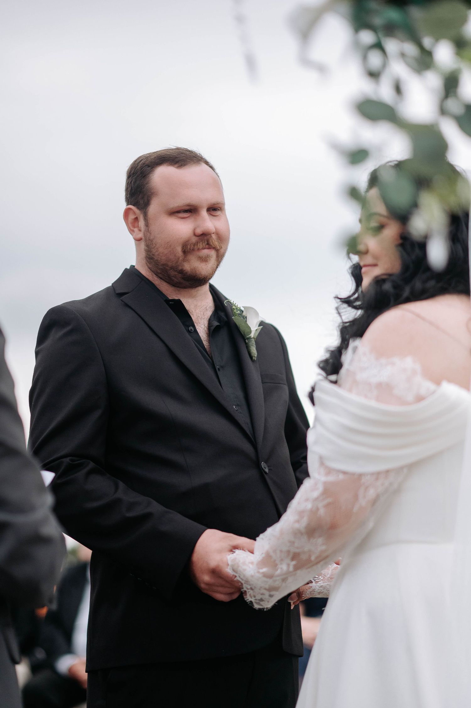 A wedding ceremony moment showing a groom in a black suit with boutonniere during the vows exchange.