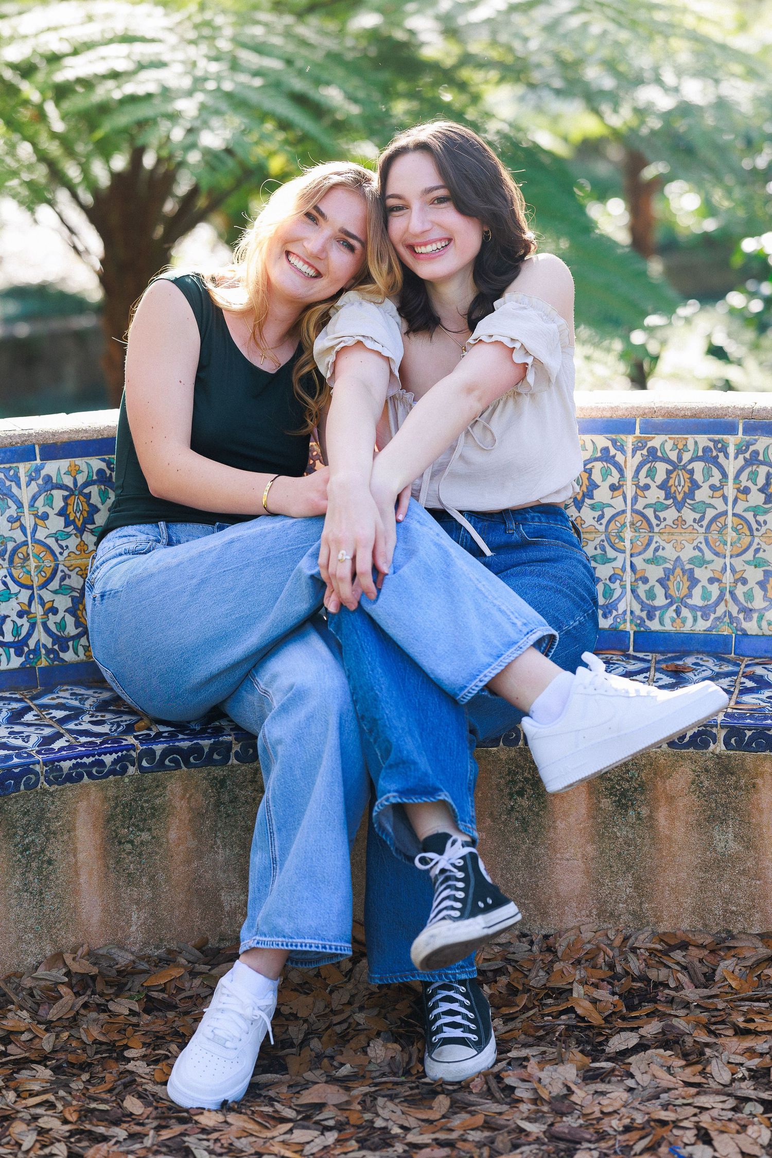 Two friends sharing a joyful moment while sitting on a decorative tiled bench in a garden setting.