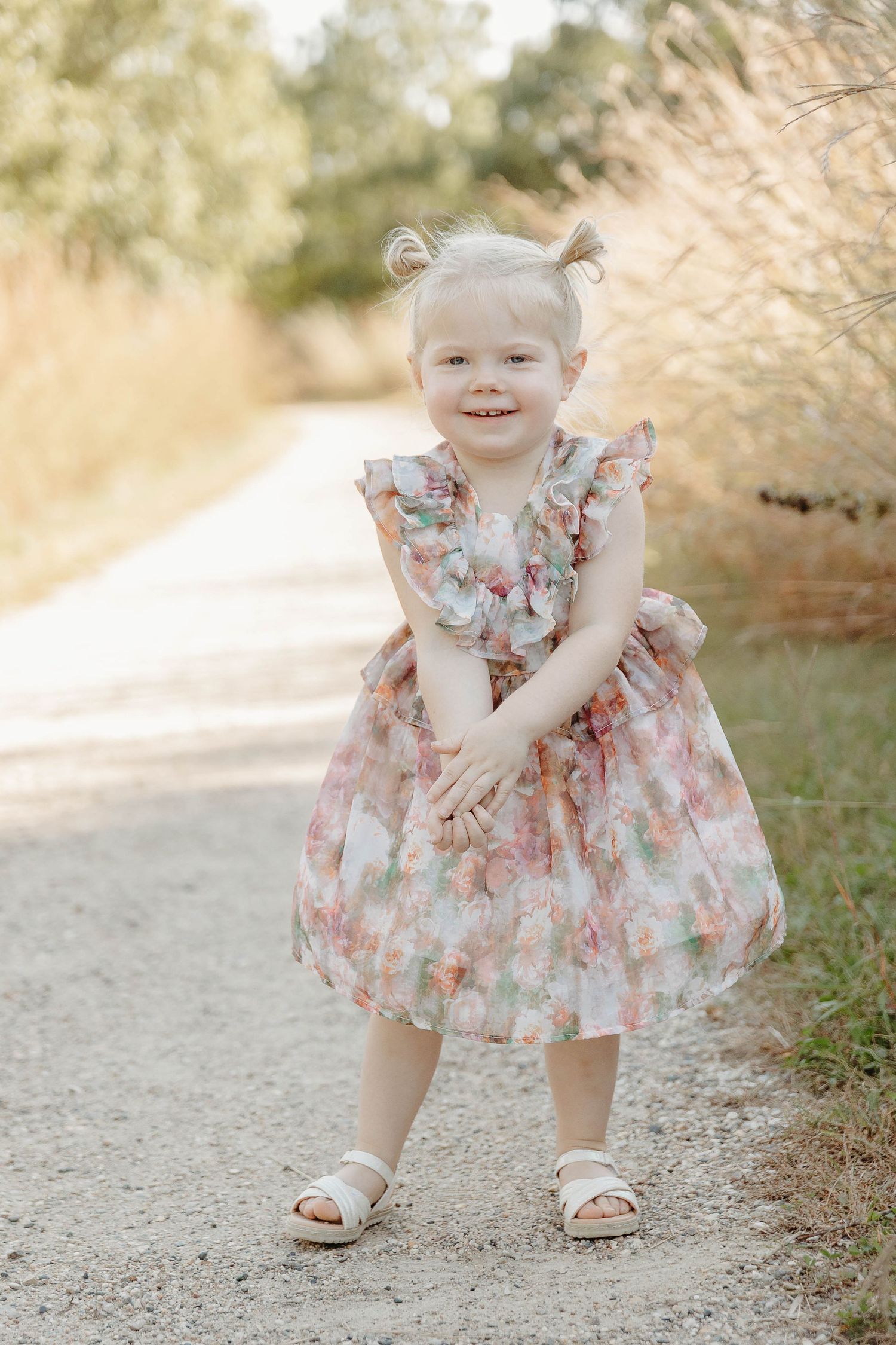 A young child in a floral dress bends down to play outdoors on a dirt path in a natural setting.