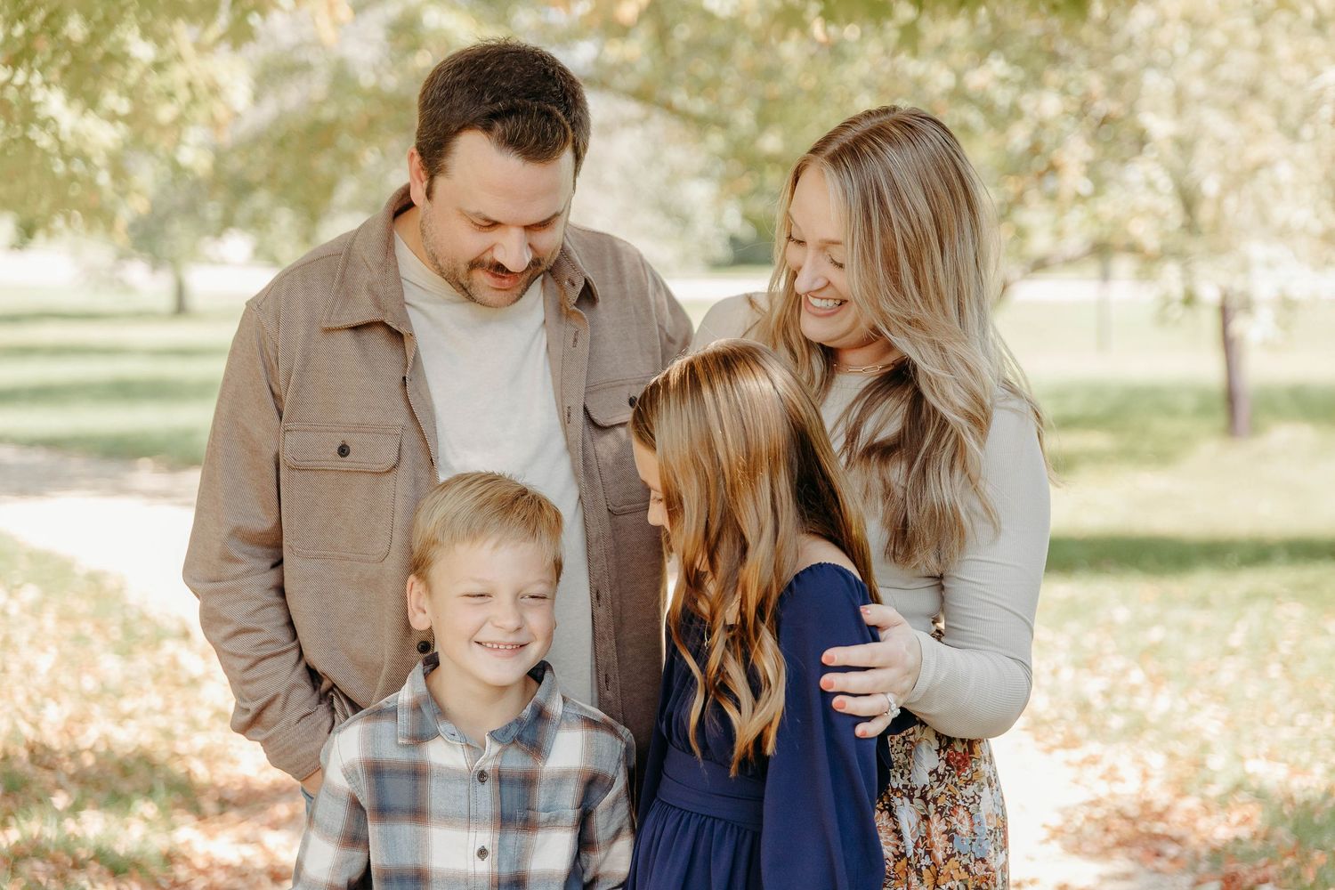 Family wearing coordinated blue and tan outfits embraces while laughing together in a sunlit outdoor setting.