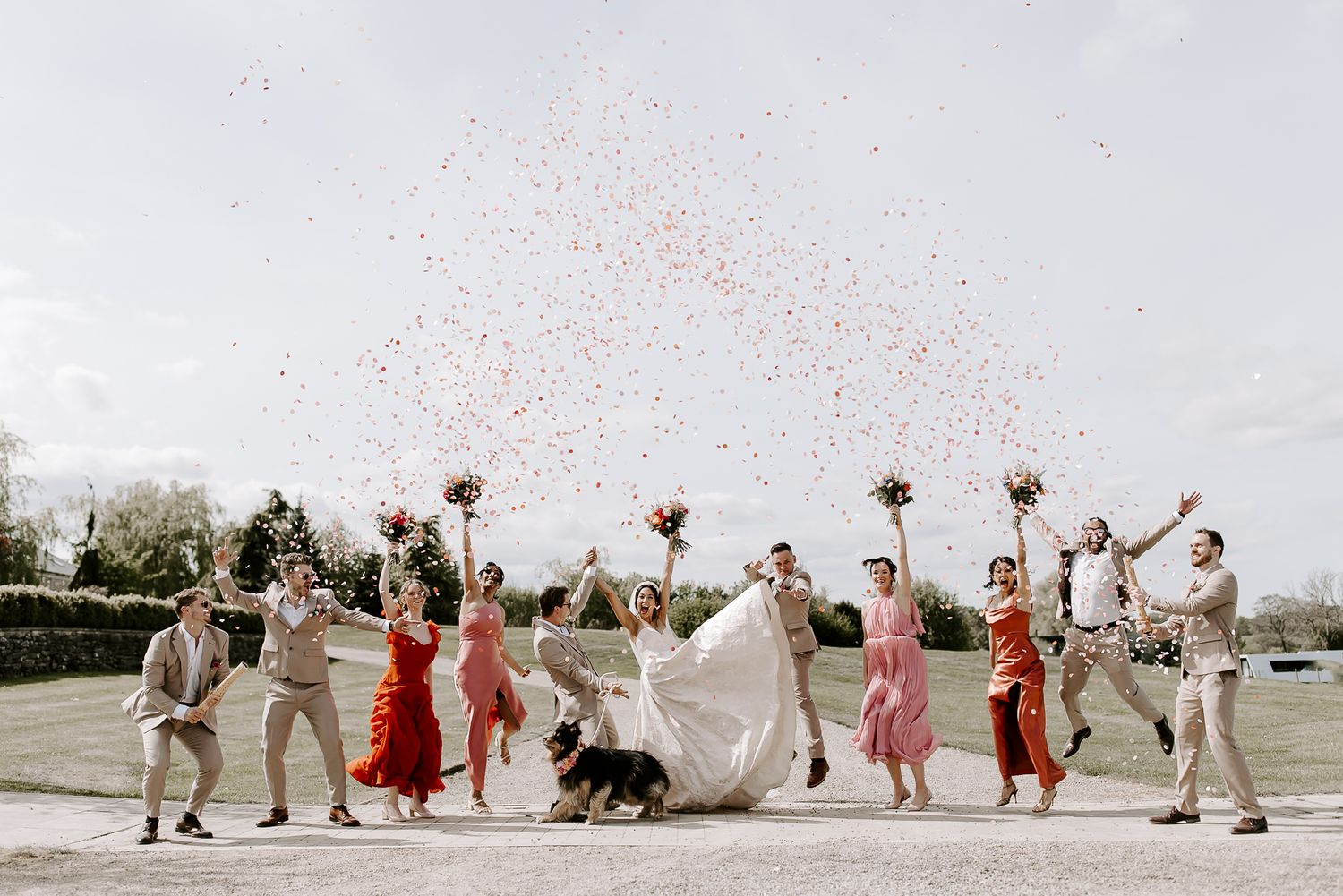 Wedding party jumps joyfully in a line against a bright natural outdoor setting wearing mix of formal attire.