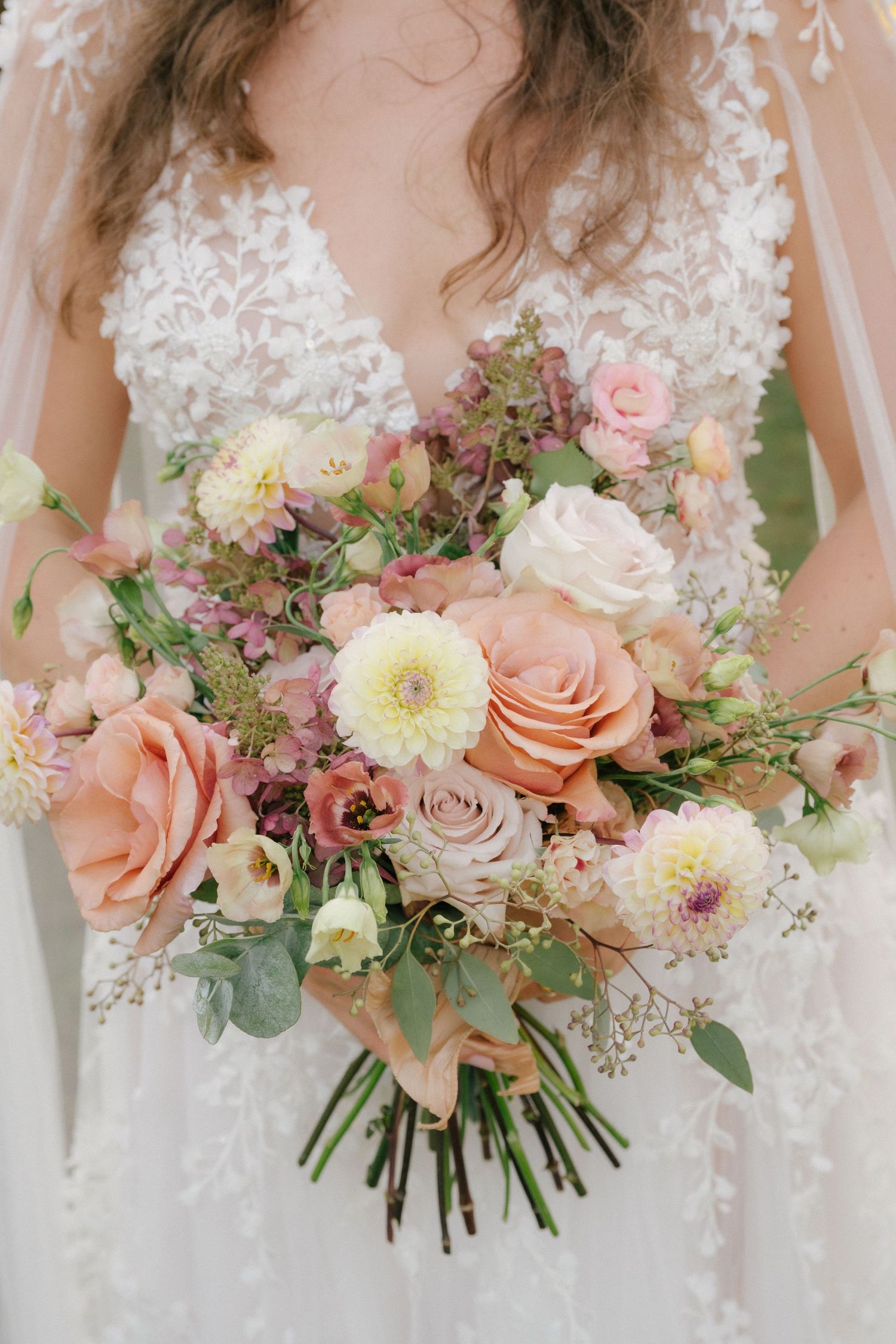 Elegante bouquet da sposa con rose color pesca, dalie bianche e vegetazione di eucalipto disposti in stile romantico.
