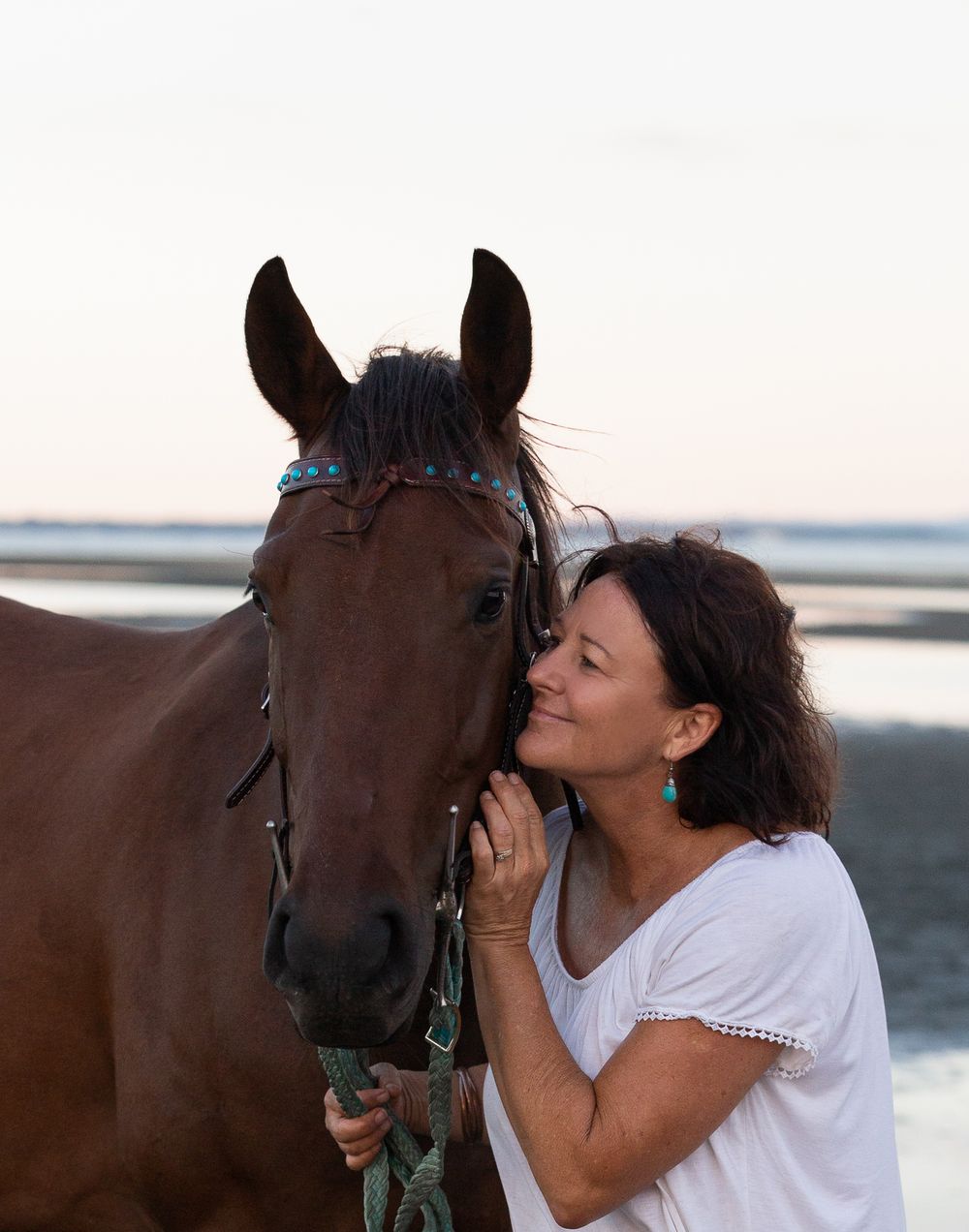 Affectionate moment between a person and their brown horse on the beach at sunset.