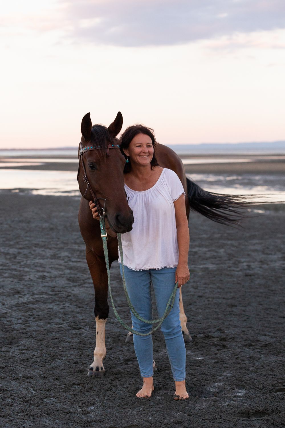 Person in white shirt and jeans sharing a loving moment with their horse on the beach.