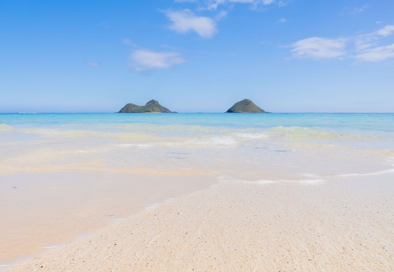 Pristine white sand beach with crystal clear turquoise water and two small tropical islands on the horizon under blue sky.