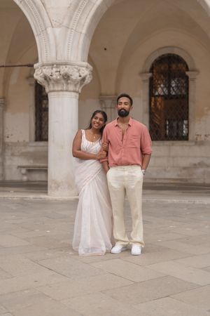 A couple in coordinated pink and white outfits poses romantically under ornate stone archways in Venice.