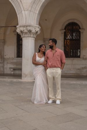 A couple poses romantically in elegant attire against classical architecture with ornate columns and arches in Venice.