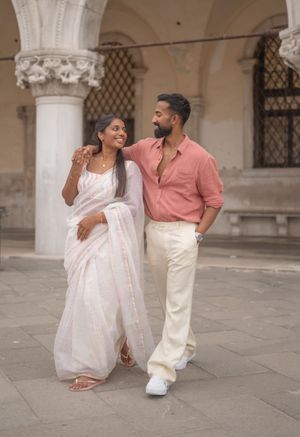A couple shares a joyful moment in traditional Indian and modern fusion attire against an ornate architectural backdrop.