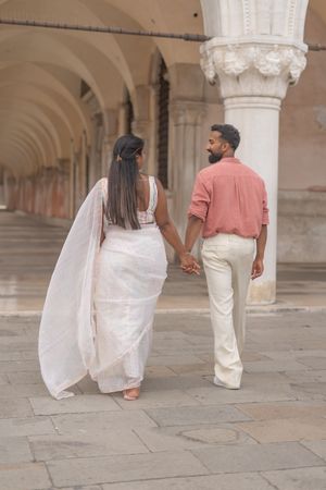 Walking couple in pink and white attire photographed from behind along Venice's historic stone colonnade.