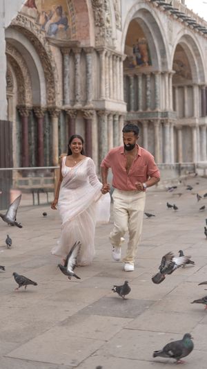 A couple walks through St. Mark's Square in Venice surrounded by pigeons and ornate Gothic architecture.