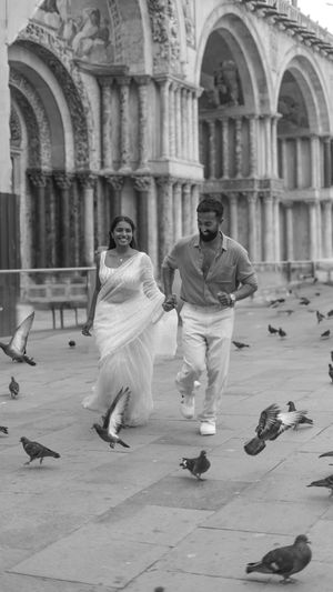 A romantic couple runs through St. Mark's Square in Venice as pigeons scatter around them in a black and white photograph.