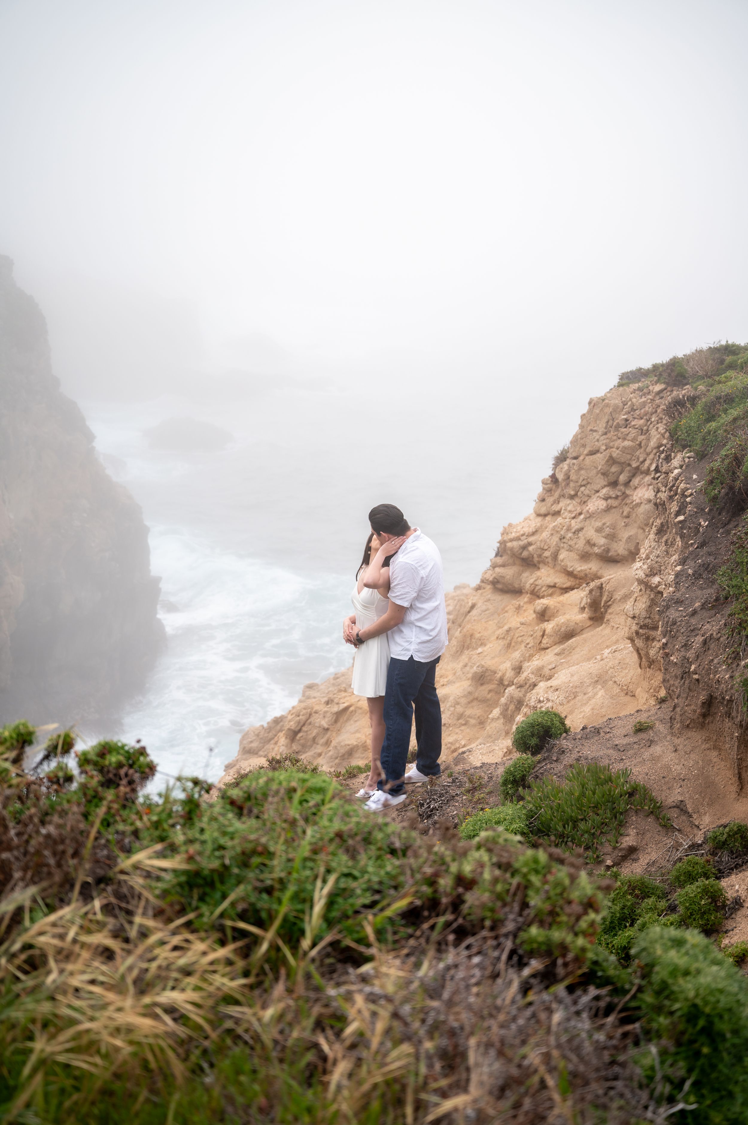 A romantic couple embraces on a misty California coastal cliff overlooking dramatic ocean views.