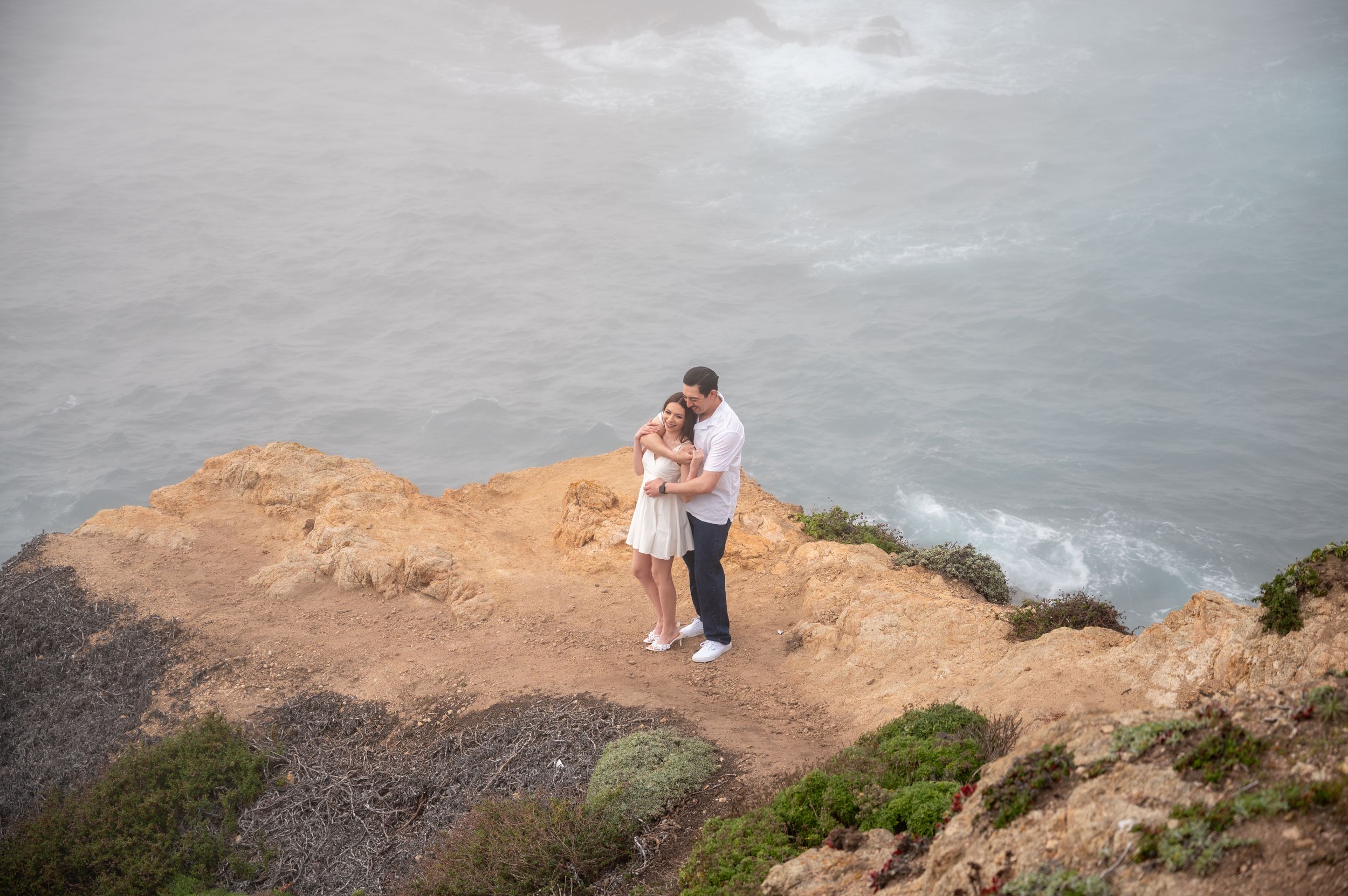 A couple embraces on a dramatic coastal cliff overlooking a misty ocean vista during an overcast day.