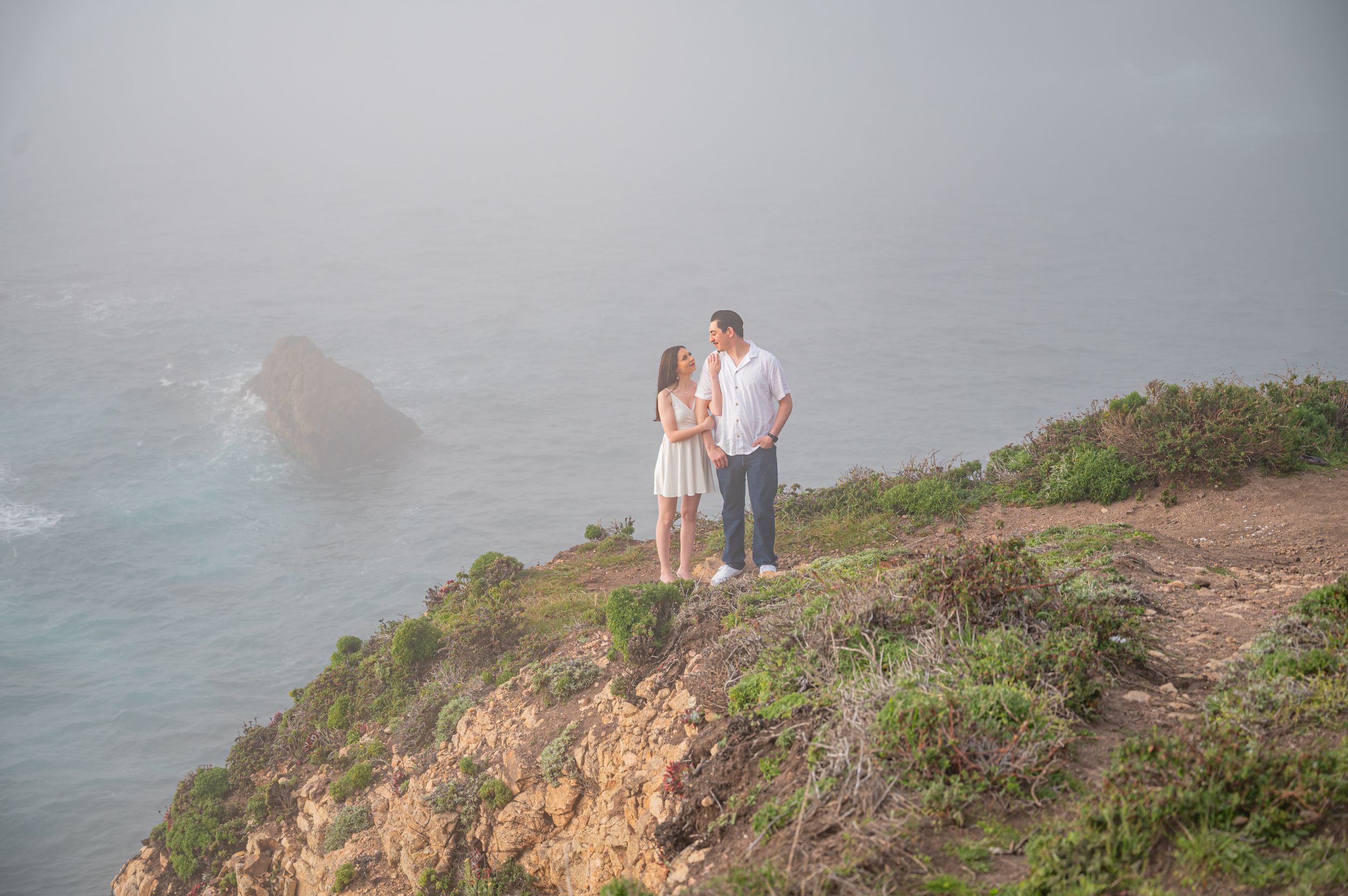 Couple stands together on rugged coastal cliffs overlooking the misty Pacific Ocean at sunset.
