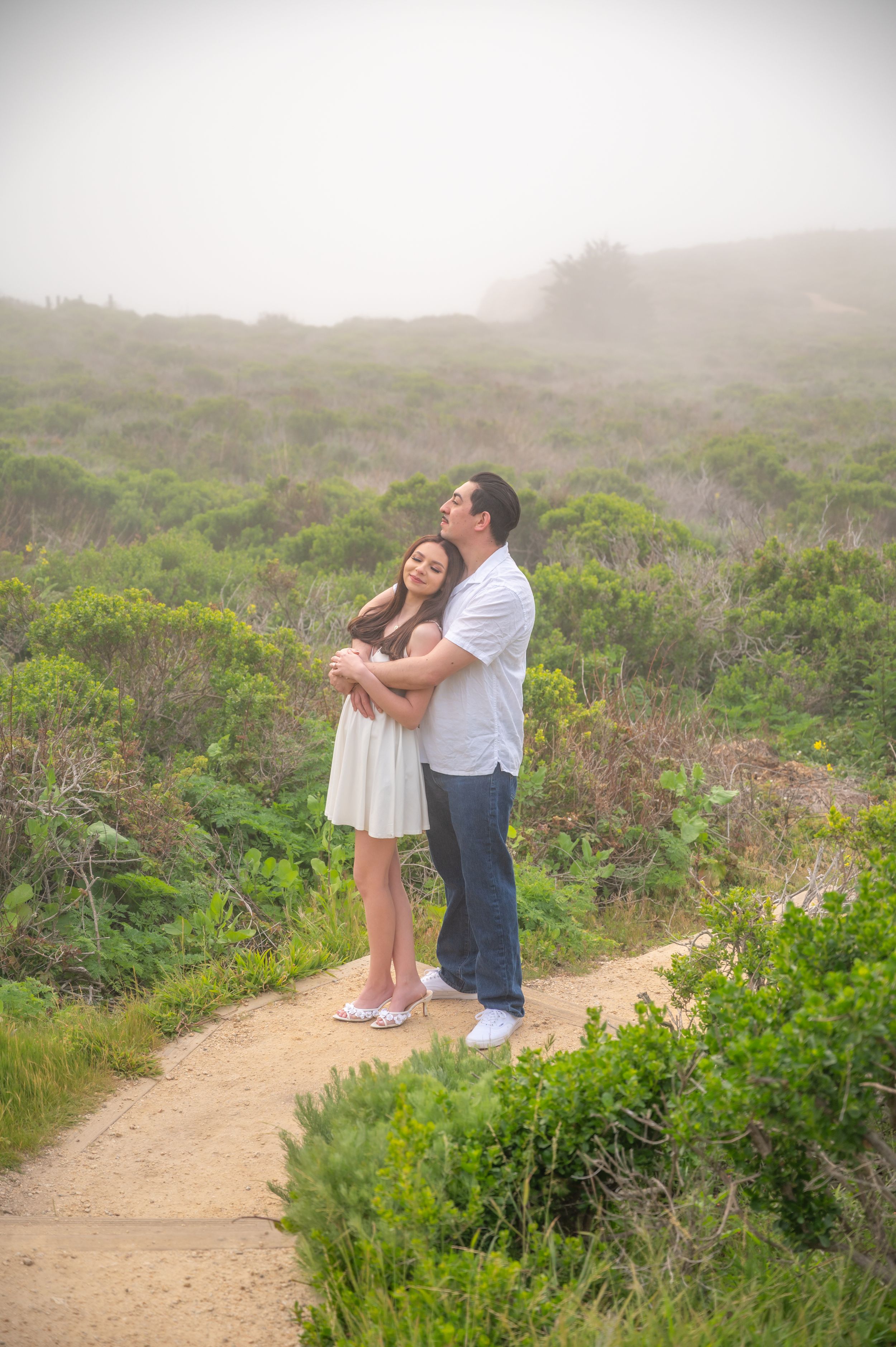 A couple shares a tender moment on a misty hiking trail surrounded by coastal vegetation on a foggy day.