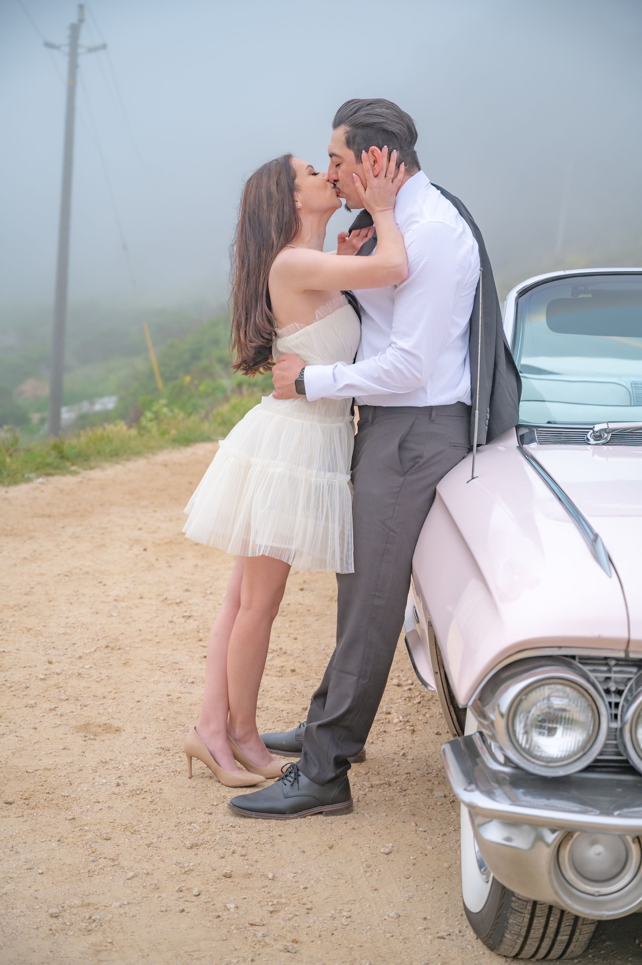 A couple shares a romantic kiss next to a vintage car on a misty dirt road.