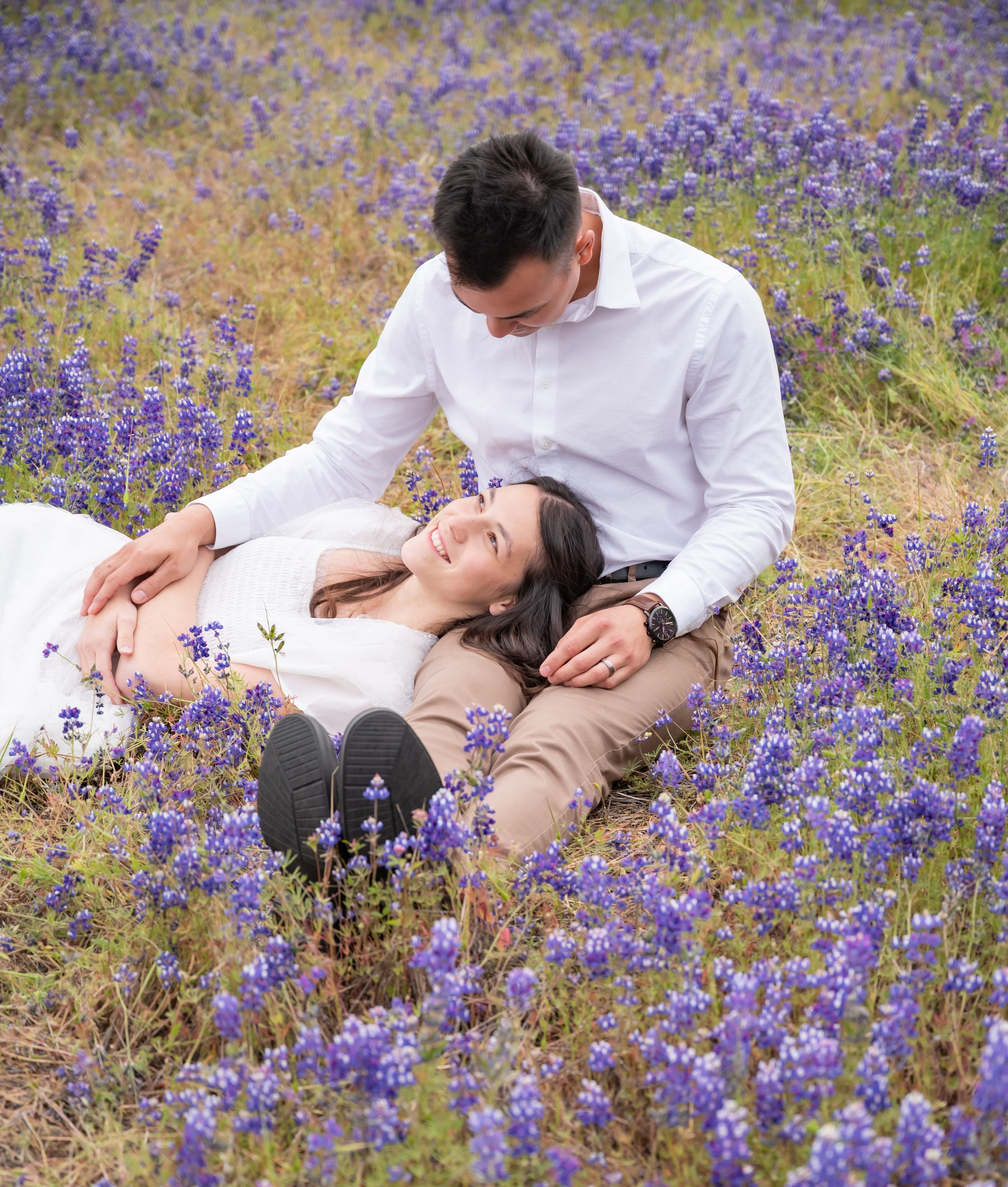A couple lies together in a blooming field of purple wildflowers on a warm sunny day.