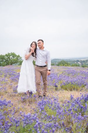 A couple stands together in a field of blooming purple wildflowers on a misty spring day.