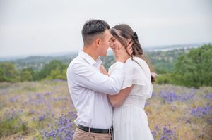 A romantic couple embraces in a field of purple wildflowers on a hilltop with a scenic valley view in the background.