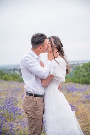 A romantic wedding couple embraces in a field of purple wildflowers with a scenic overlook in the background.