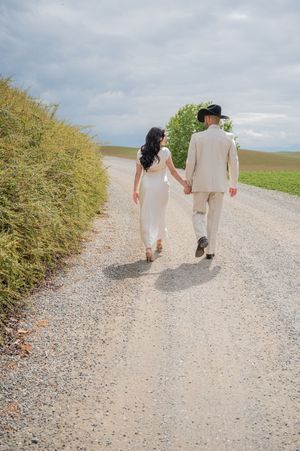 Couple walks hand in hand down a gravel path surrounded by yellow brush on a sunny day.