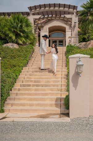 A pair stands elegantly on grand stone steps leading to an archway entrance framed by palm trees and Tuscan architecture.