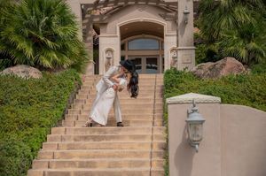 Couple share romantic dip kiss on elegant stone staircase surrounded by lush greenery and palm trees at grand entrance.