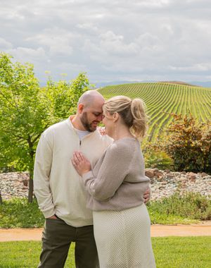 A couple shares an intimate moment in front of scenic vineyard rows under cloudy skies.