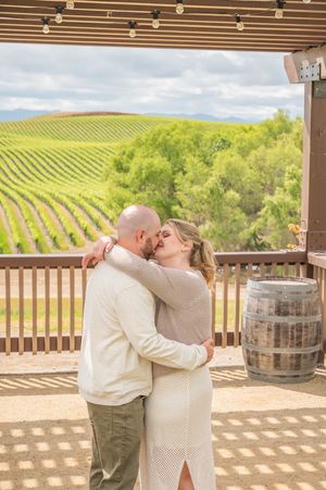 Couple embraces on a winery deck overlooking rows of grapevines on rolling hills under cloudy skies.