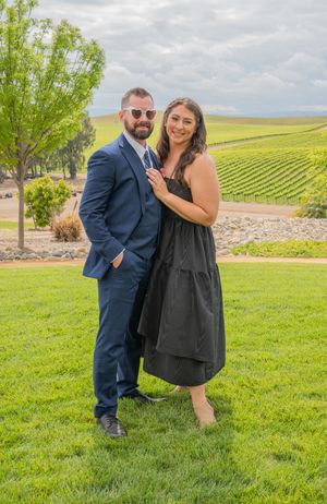 A couple poses playfully on green grass with rolling vineyard hills stretching into the distance.