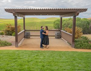 A couple embraces under a wooden pergola overlooking scenic vineyard hills and green fields in wine country.