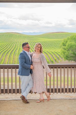 A couple shares a romantic moment on a winery deck overlooking rolling vineyard hills on a partly cloudy day.