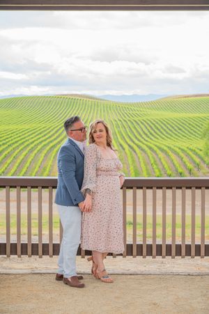 Two figures stand close together overlooking rolling green vineyard hills on a cloudy day.