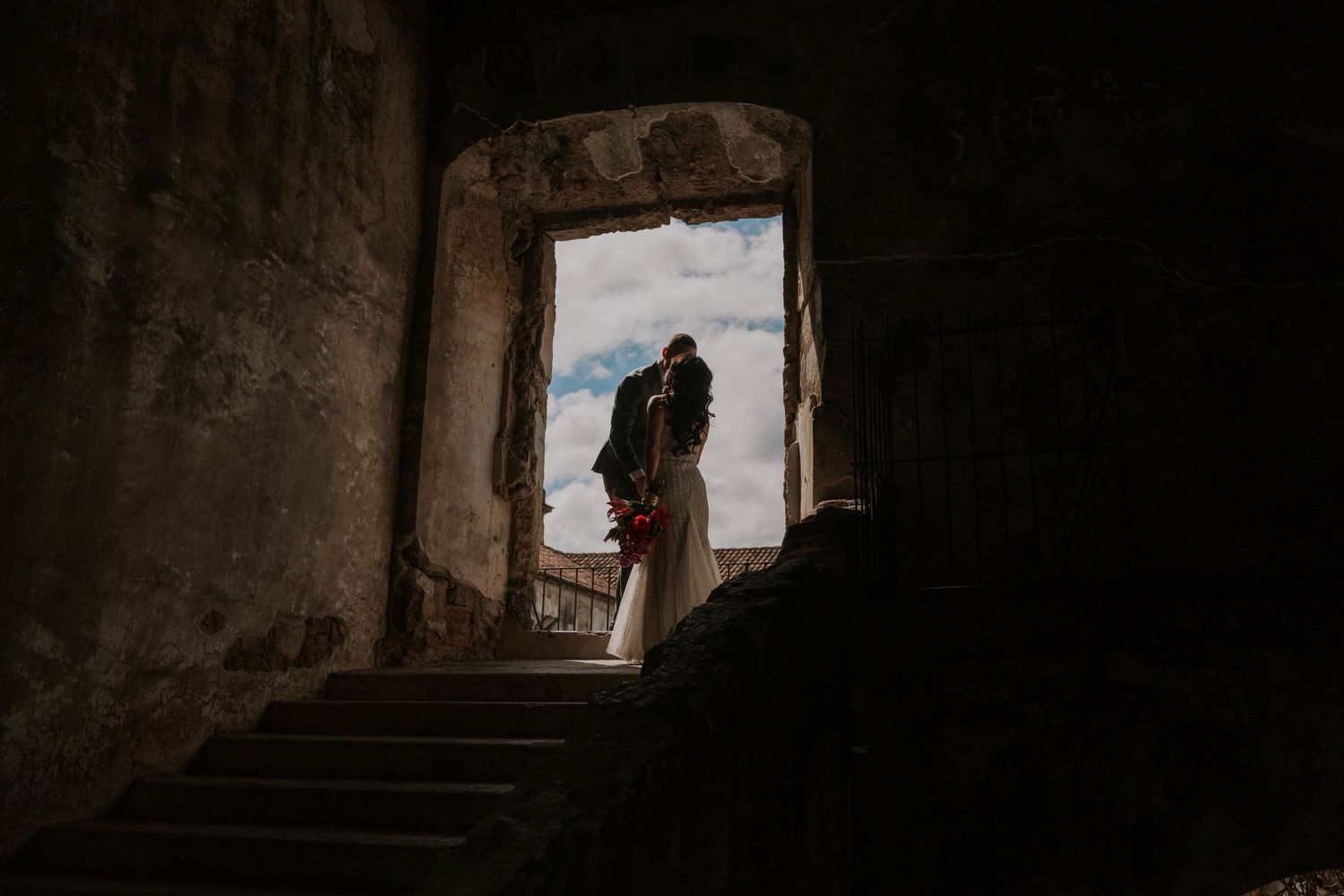 Silhouetted figure stands in stone doorway at top of dark staircase overlooking cloudy landscape.