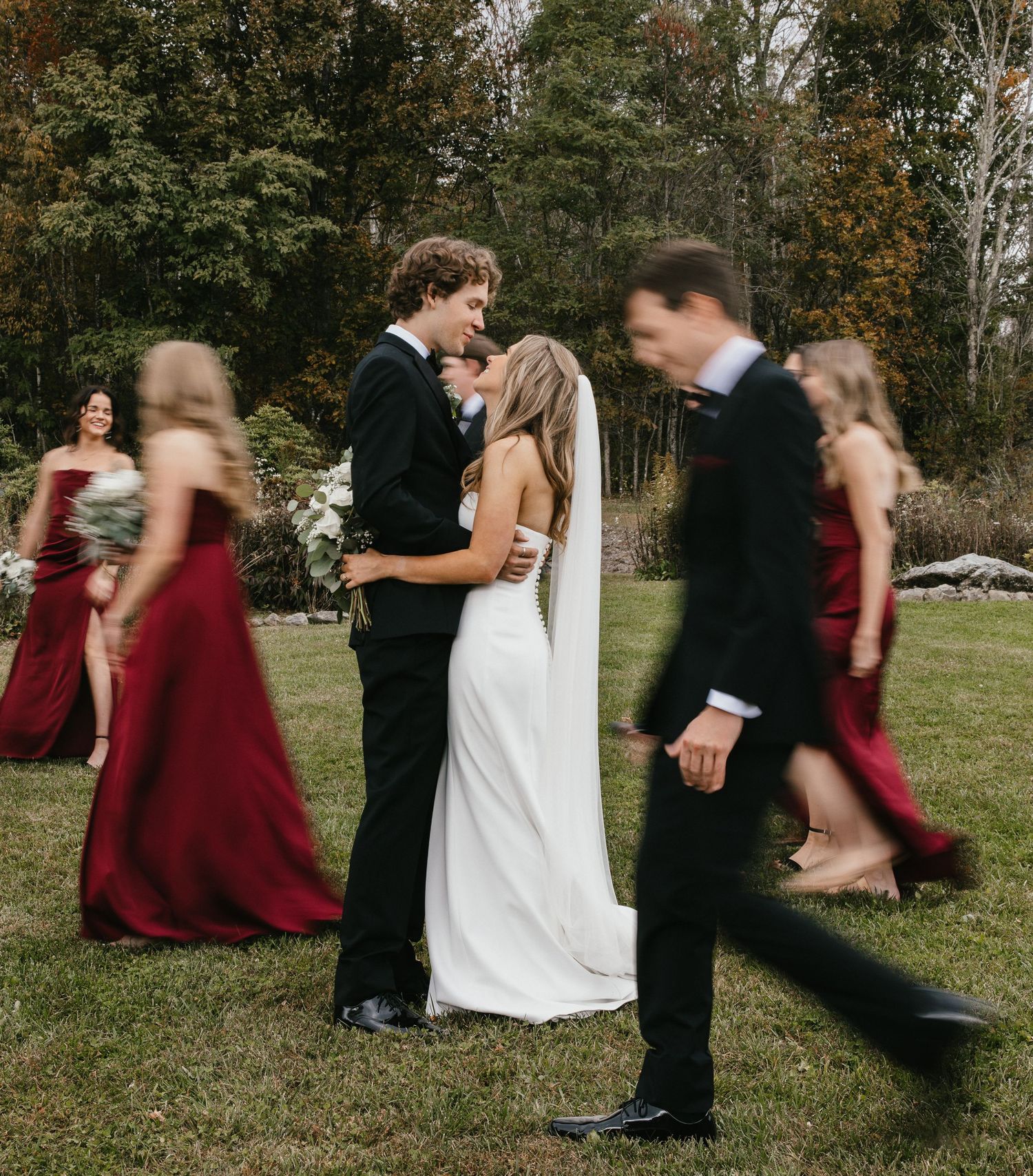 Wedding party members in burgundy dresses and black suits dance in a playful sequence on a grassy lawn.