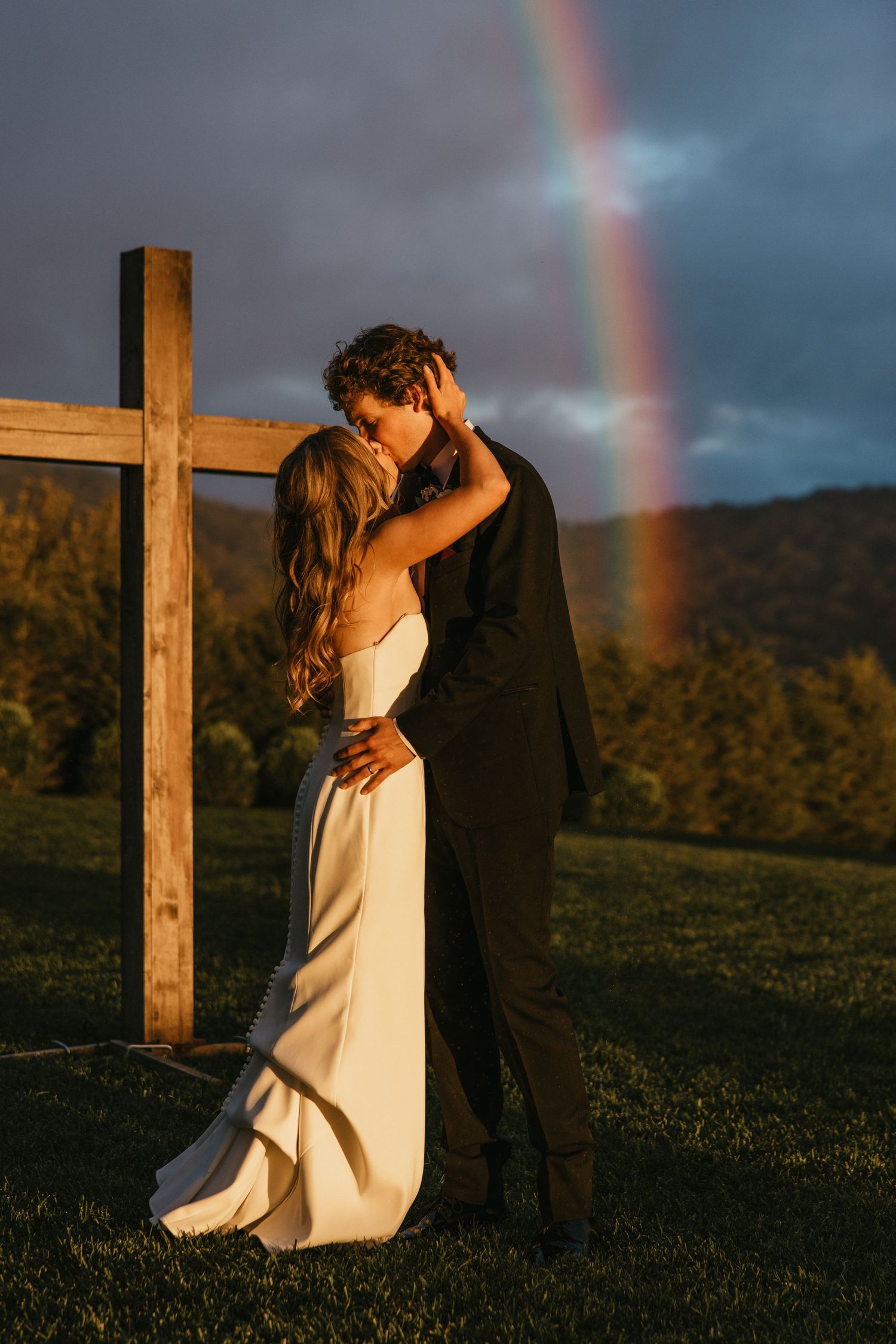 A couple embraces on a grassy hillside with a vibrant rainbow arching across the dramatic stormy sky in the background.