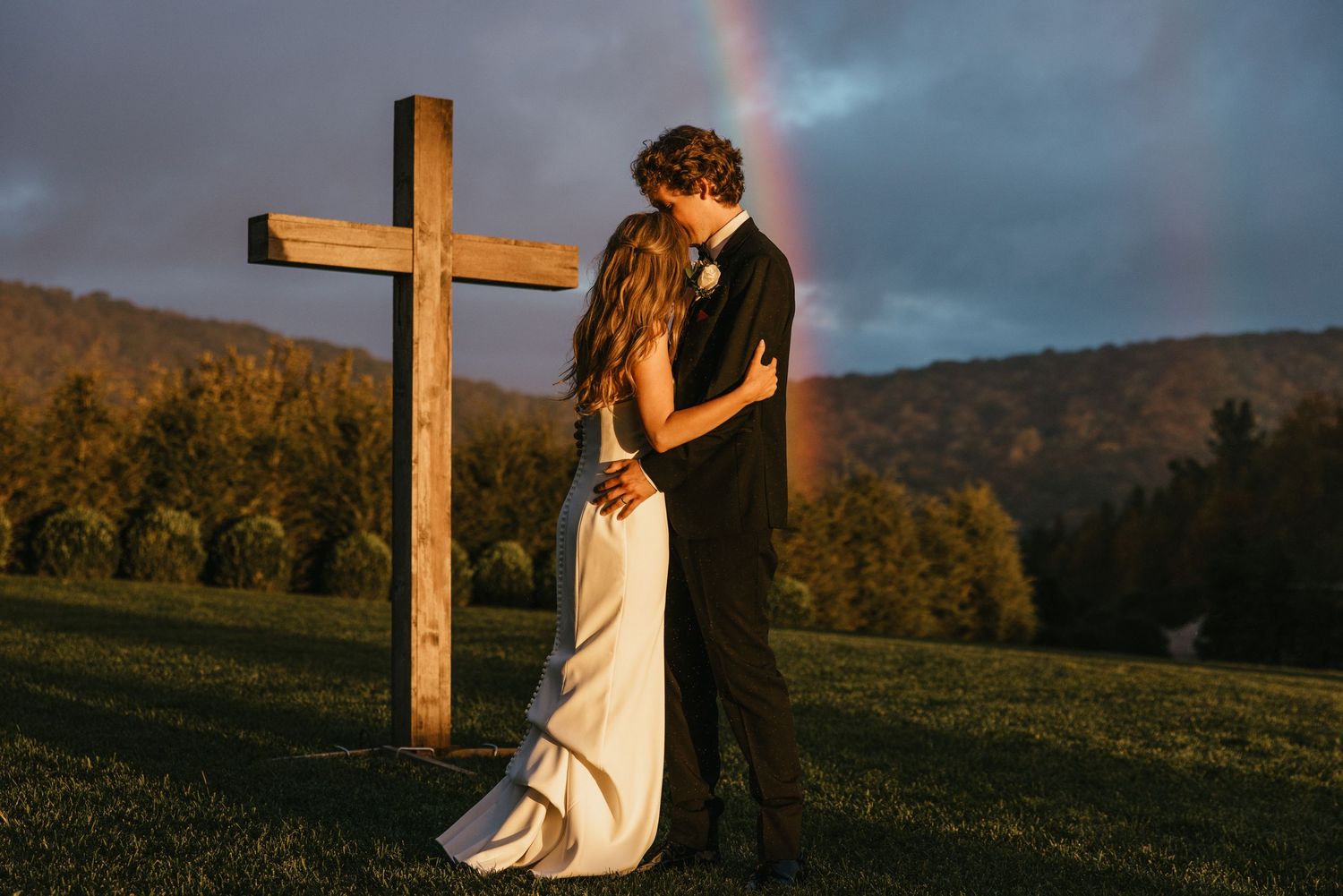 A wooden cross stands silhouetted against a dramatic cloudy sky with a rainbow in the background.