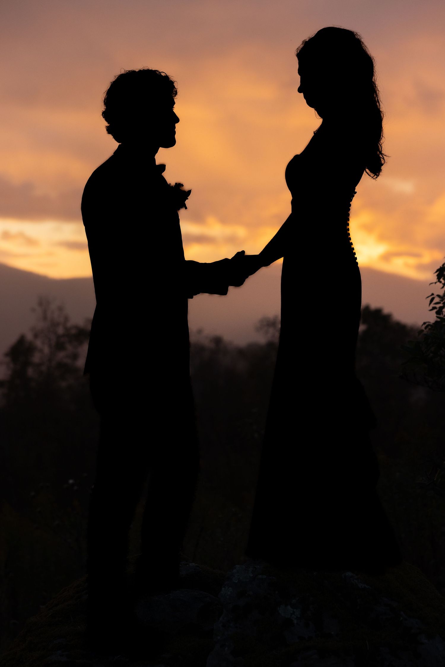 Silhouettes of two people against a dramatic orange sunset sky with dark trees in the background.