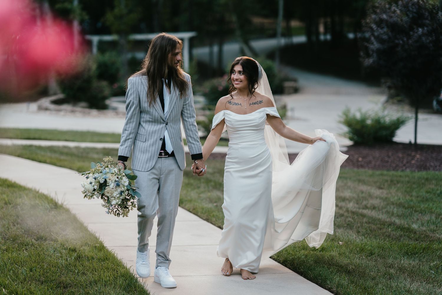 A couple in wedding attire walks together on a path holding hands while carrying a white floral bouquet.